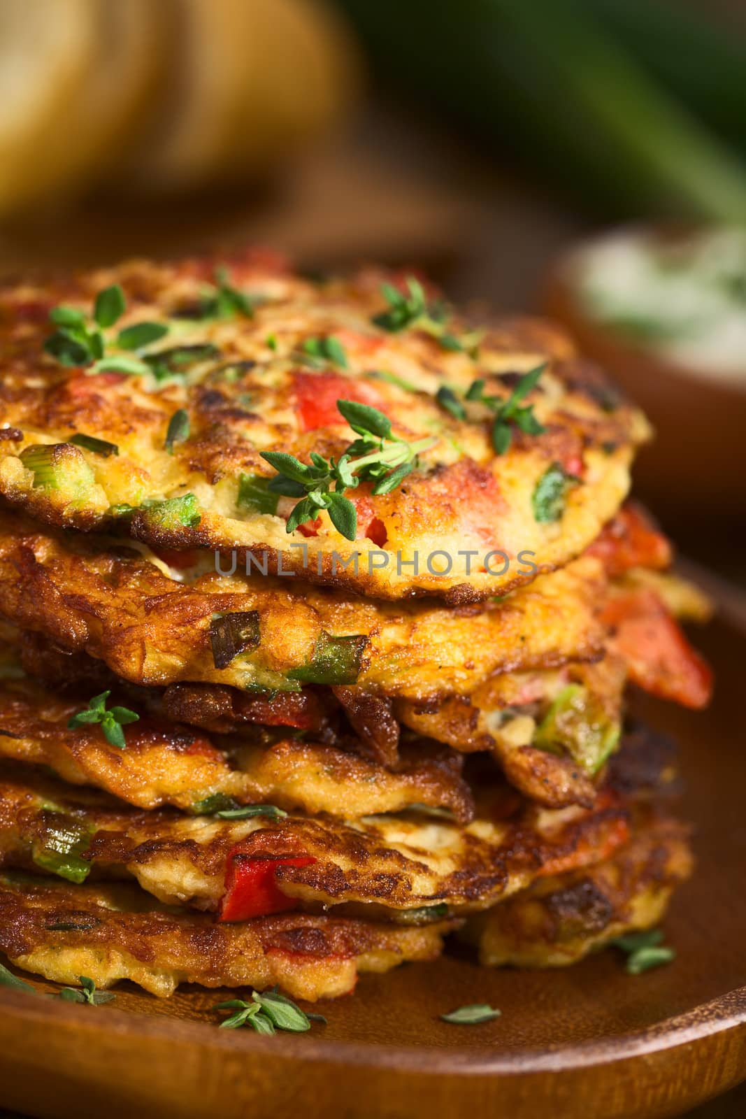 Vegetable and egg fritter made of zucchini, red bell pepper, eggs, green onions and thyme piled on a wooden plate with baguette slices and yogurt dip in the back (Selective Focus, Focus on the front of the thyme sprig on the top of the fritters and on the front of the top fritters) 