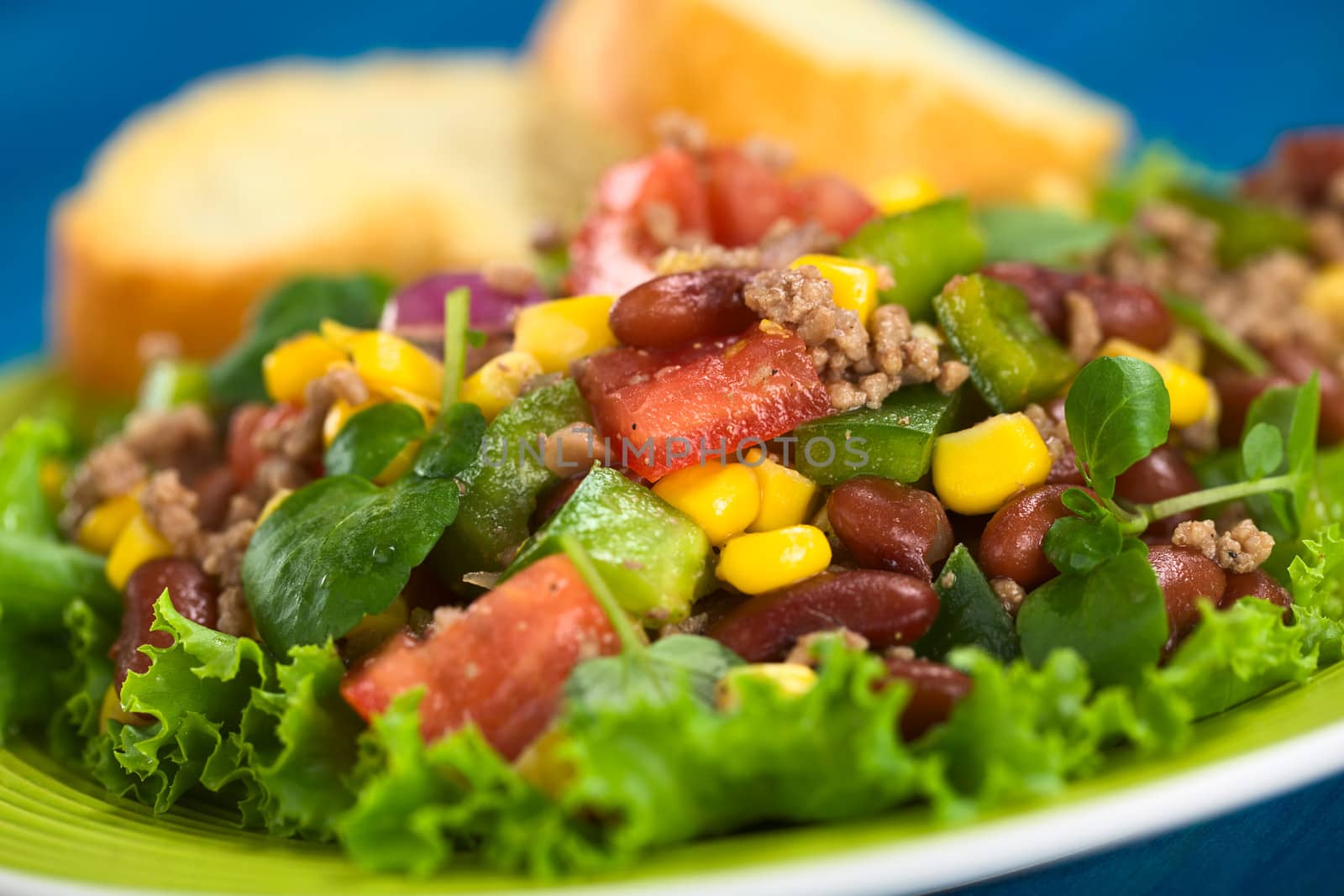 Chili con carne salad made of mincemeat, kidney beans, watercress, green bell pepper, tomato, sweet corn and red onions served on lettuce on a plate with baguette slices in the back (Selective Focus, Focus on the tomato piece in the middle of the image)