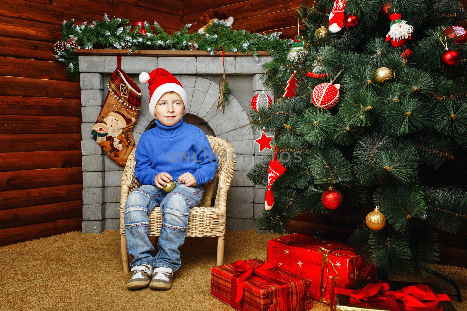 Boy in hat sitting in wicker chair near Christmas tree and gifts