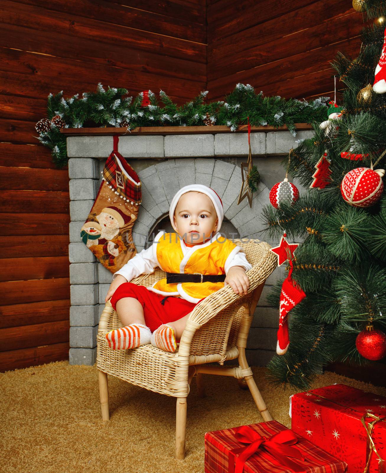 Boy in hat sitting in wicker chair near Christmas tree and gifts