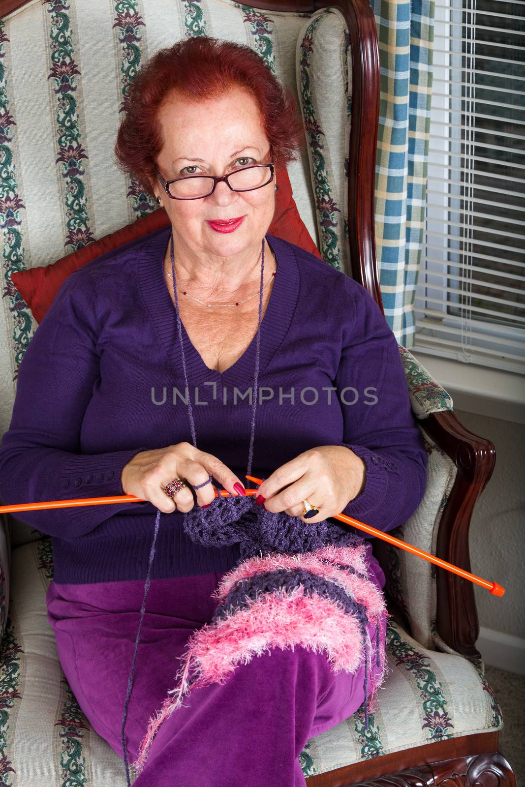 Red haired senior lady looking at you while knitting her purple scarf, she is wearing purple clothing perhaps purple is her favorite color