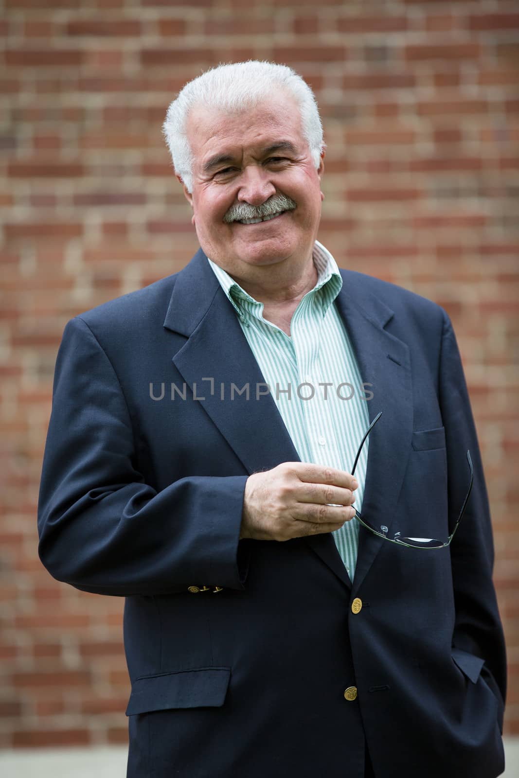 Senior adult looking at you smiling and trustfully while holding his glasses, infront of red brick wall