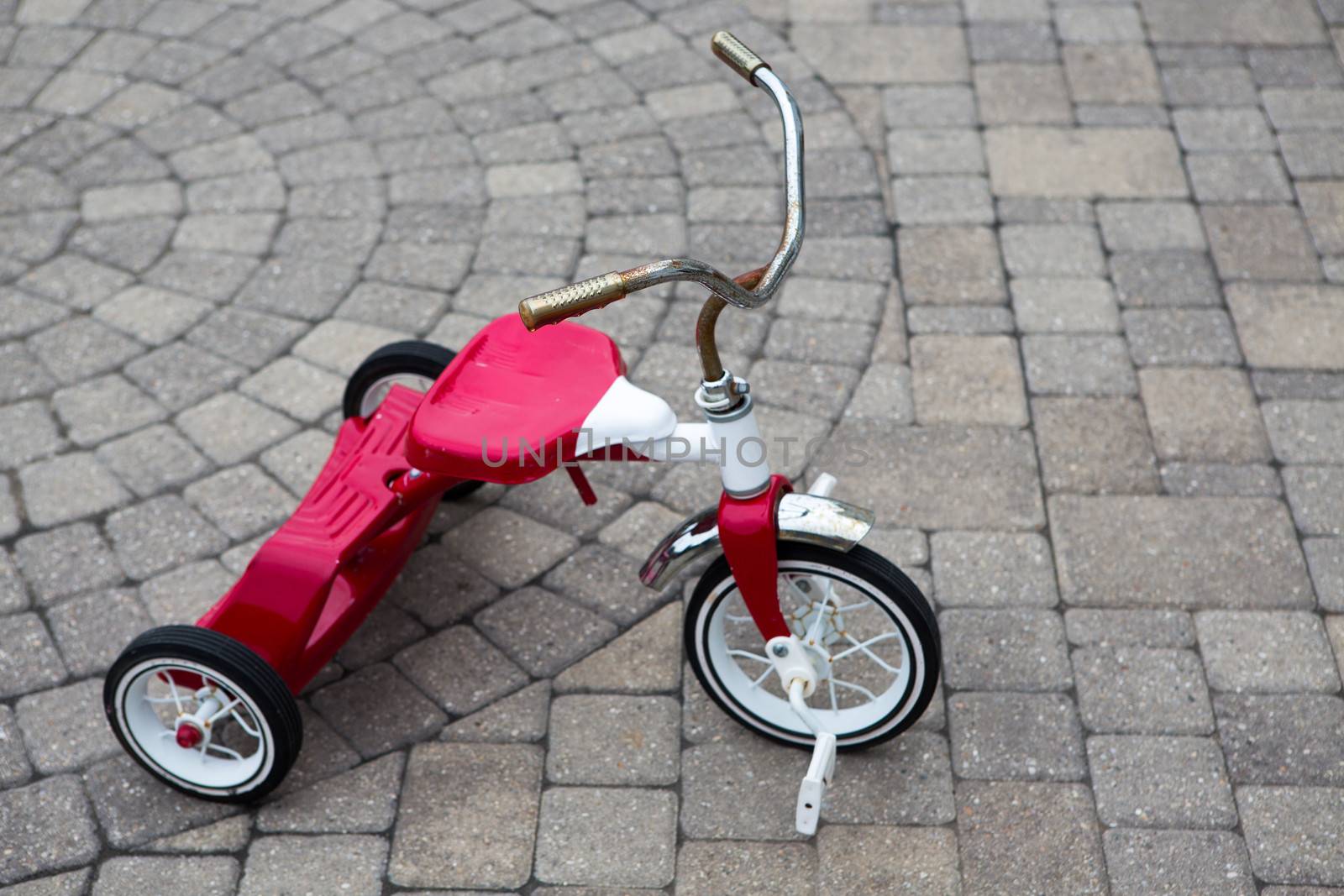 Childs red tricycle parked on a cobble design paved street