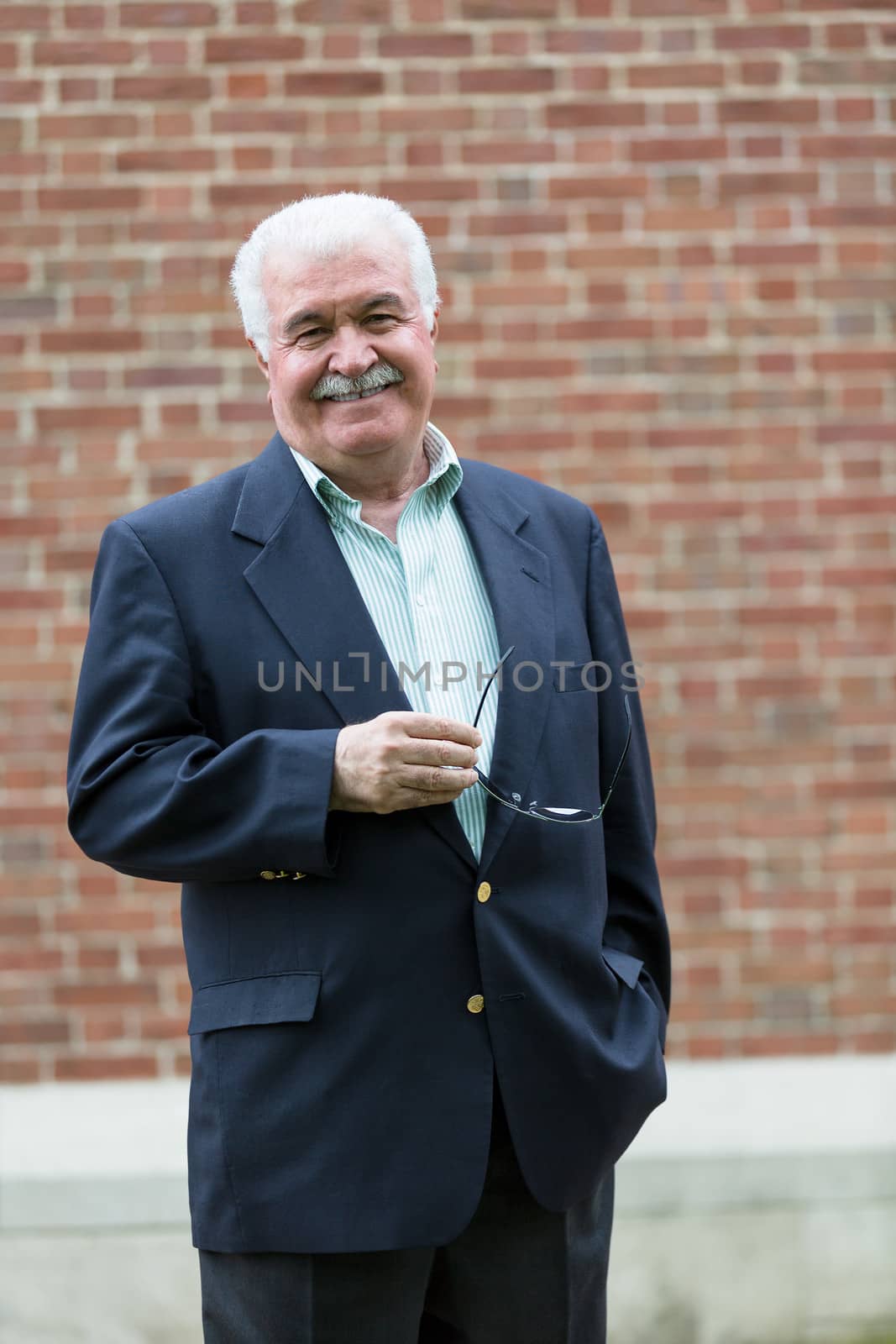 Senior adult looking at you smiling and confidingly while holding his glasses, infront of red brick wall
