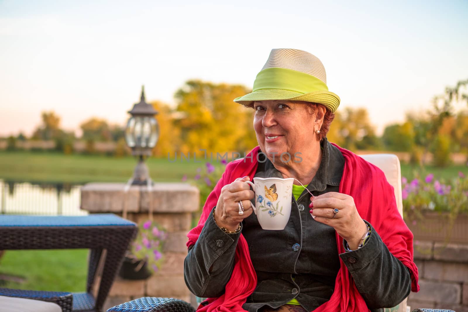 Senior woman with a straw green hat, sitting at the patio and showing her hot tea, perhaps she wants to stay warm