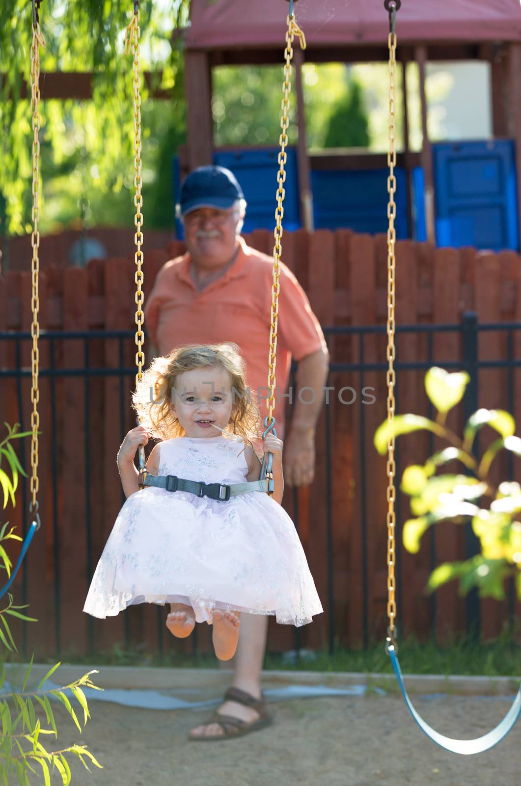Toddler girl having fun on the swing while having candy with her bare naked feet and pushed by her grandfather with the hat