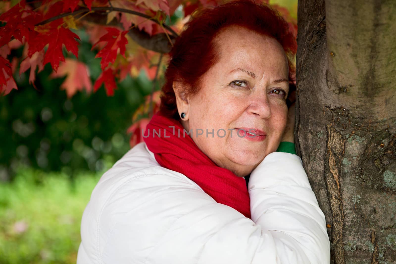 Red hair senior lady looking at you thoughtfully with her red scarf and white coat while leaning to the tree