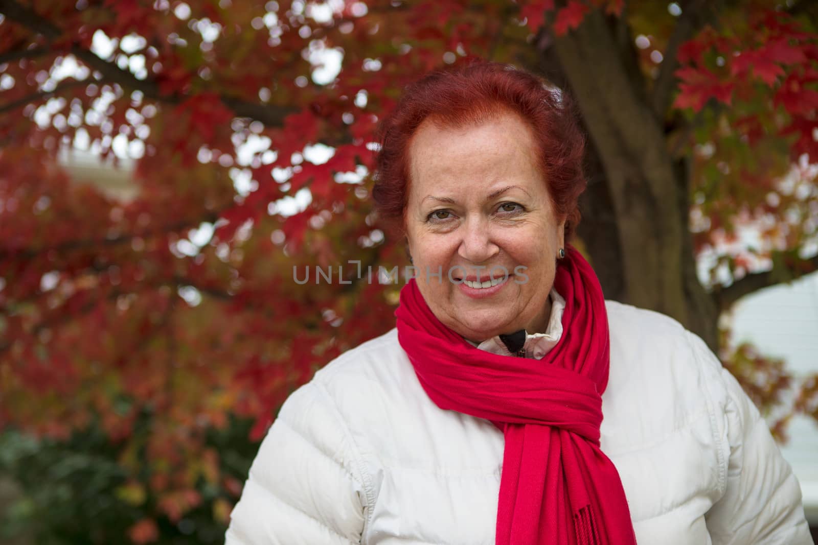 Red hair senior lady looking at you happily and trustfully with her red scarf and white coat under the tree with red falling leaves
