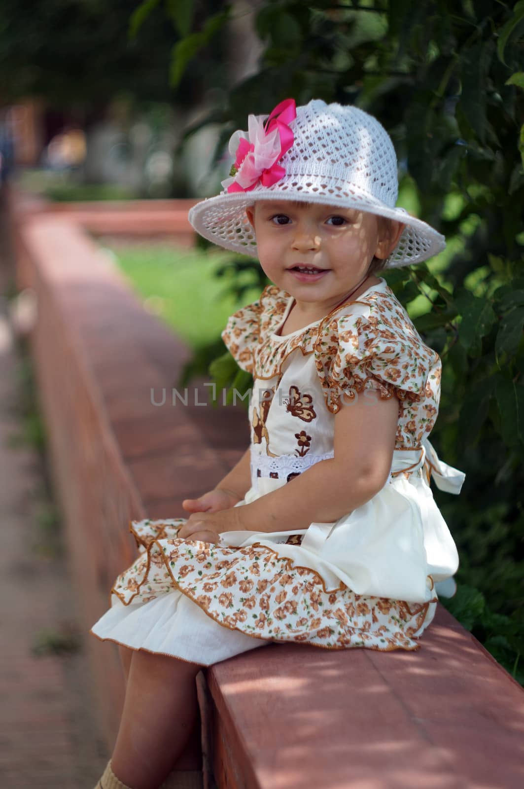 Two-year-old girl in a smart dress and hat sitting on the street                               