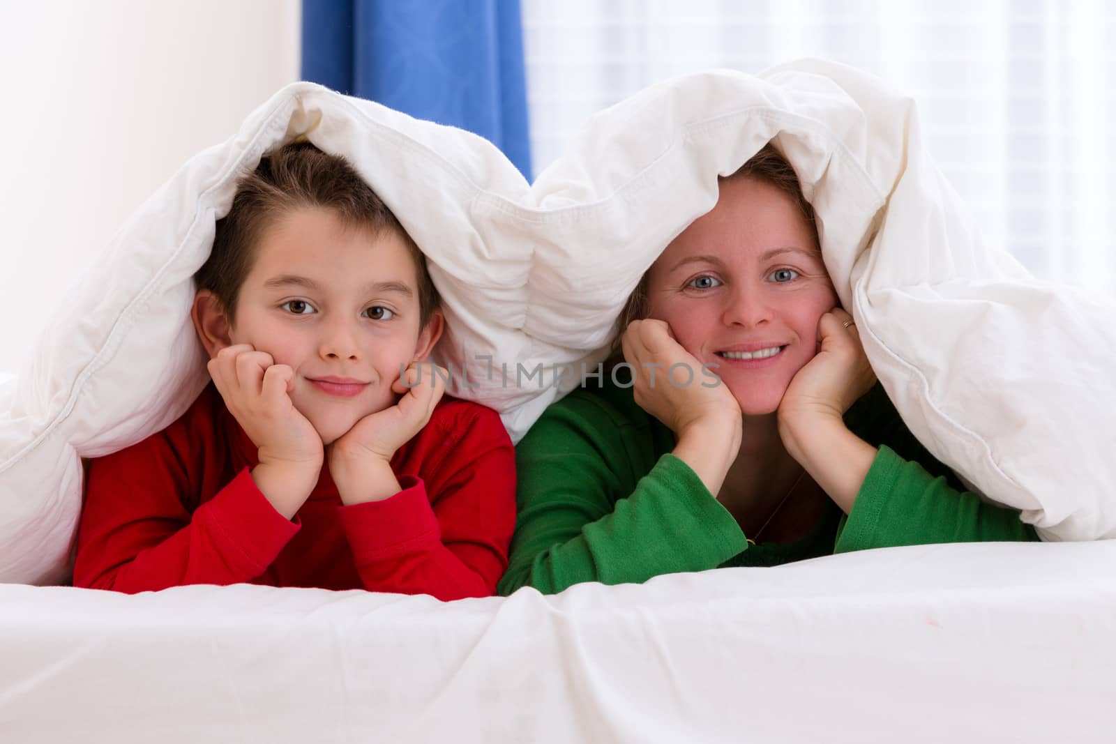 Mother and her son under the blanket with Christmas color pajamas, smiling to you