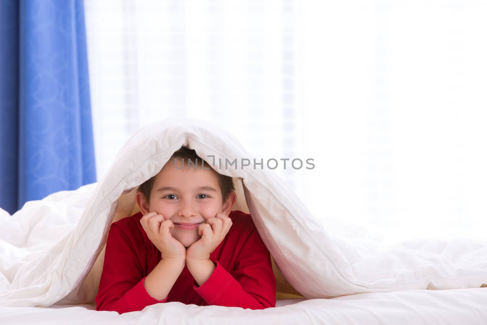 Close up portrait of a eight years old boy laying on the bed looking at  you under his warm blanket. He looks happy and relaxed.