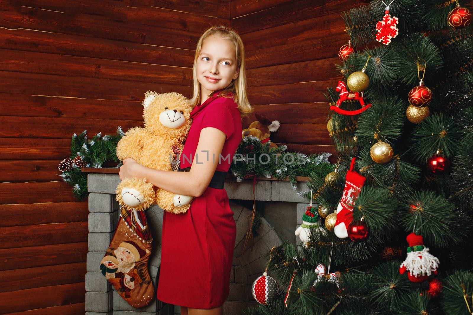 Young girl holding teddy bear and stands near Christmas tree