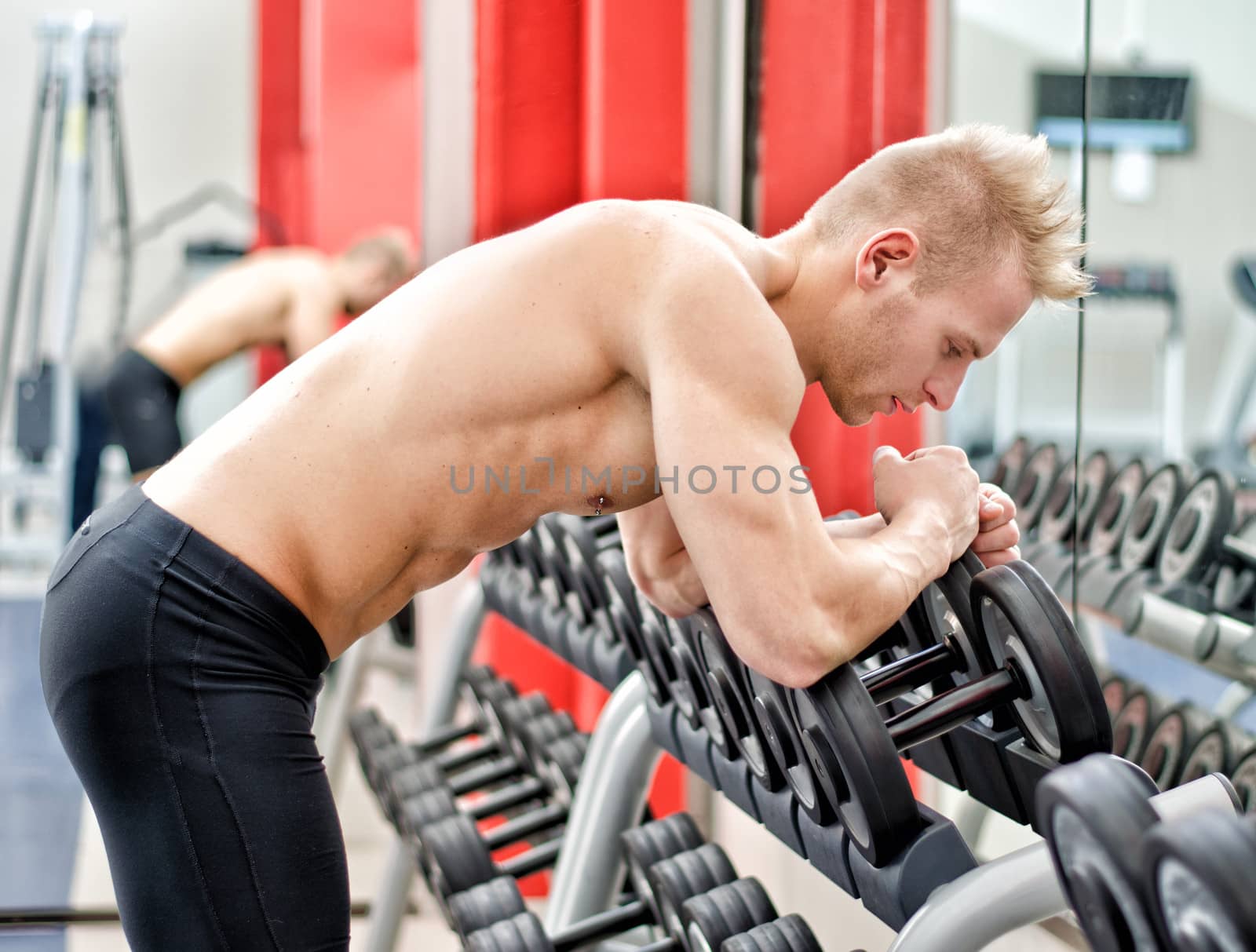 Young man resting on dumbbells rack after workout in gym by artofphoto