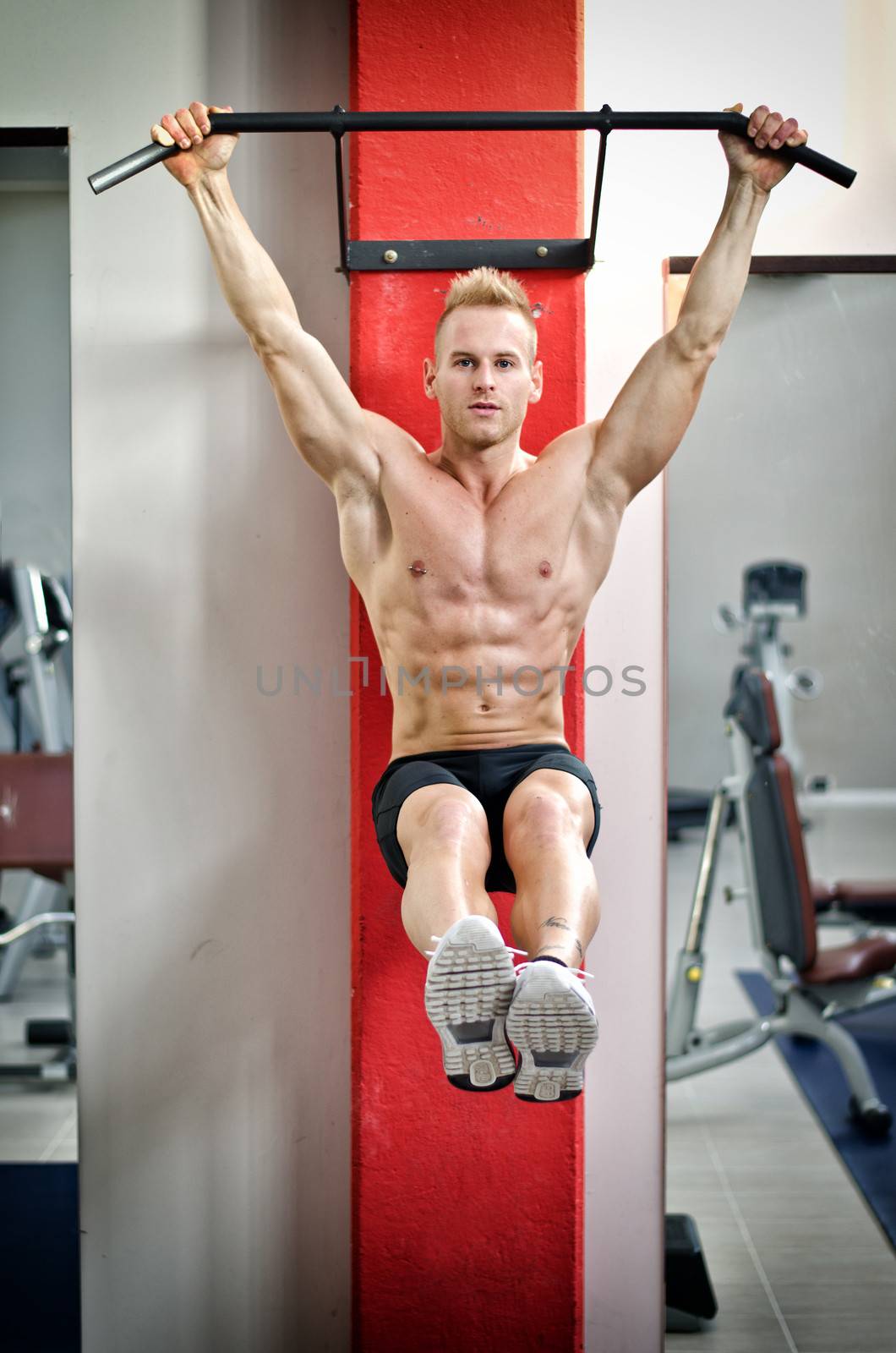 Blond, attractive young man hanging from gym equipment, looking at camera