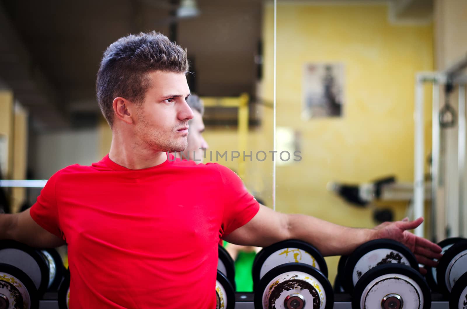 Handsome young man in gym sitting on dumbbells rack, looking to a side