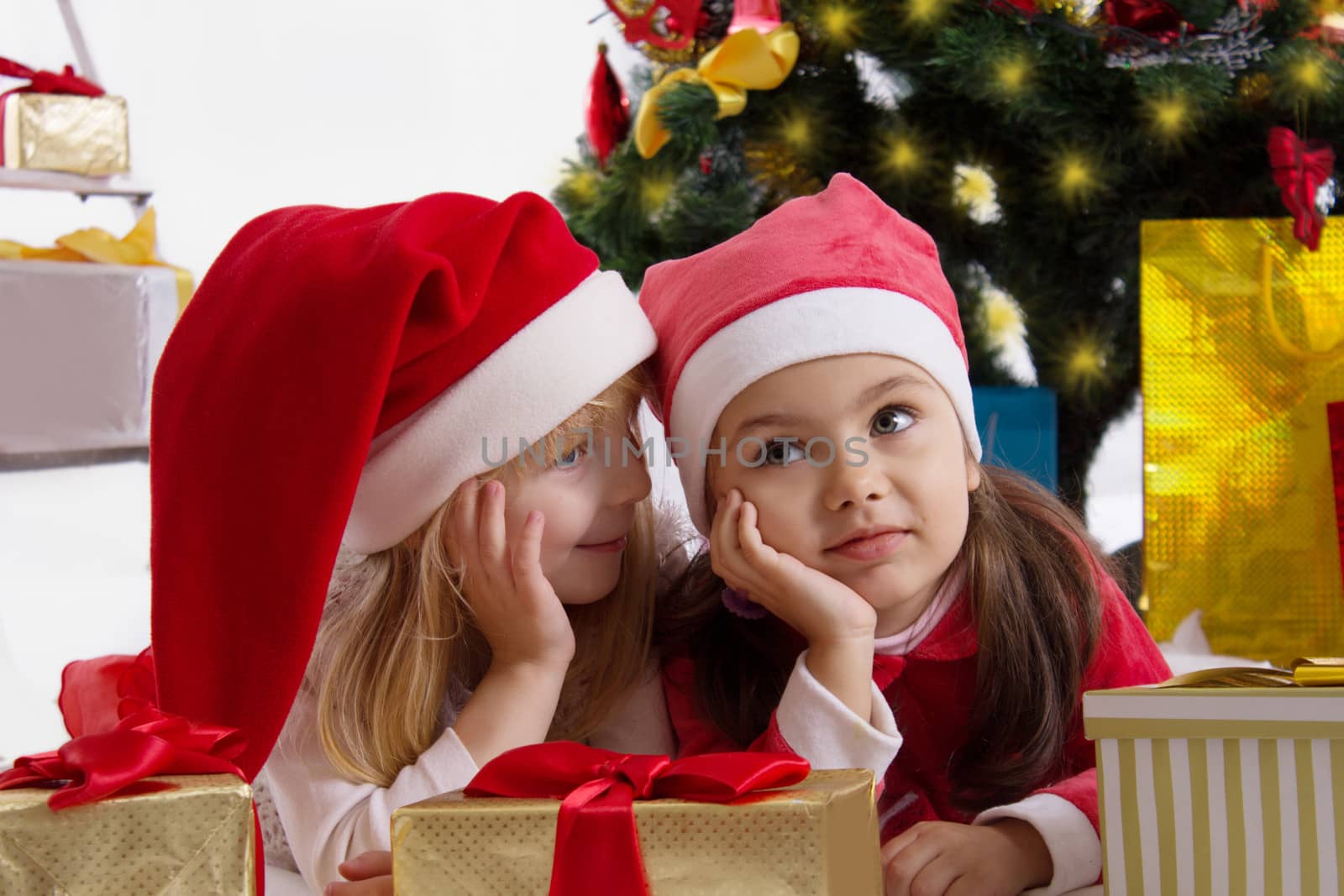Two girls in Santa hats sharing secrets under Christmas tree