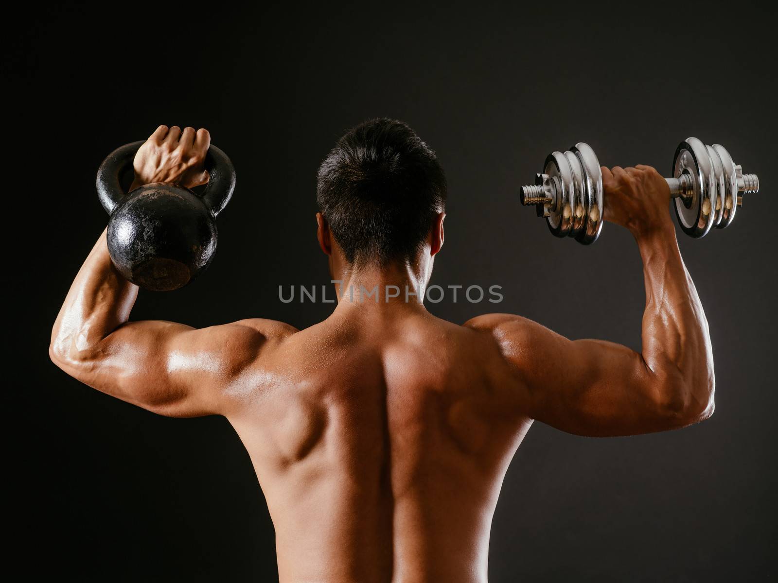 Photo of an Asian male exercising with both a kettlebell and a dumbbell, doing shoulder press over dark background.