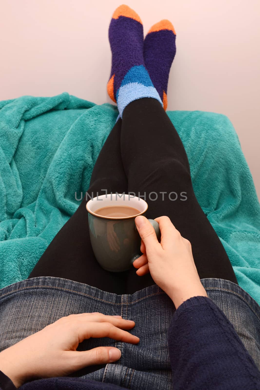 Woman dressed warmly, holding hot coffee or tea, with her feet up on the sofa