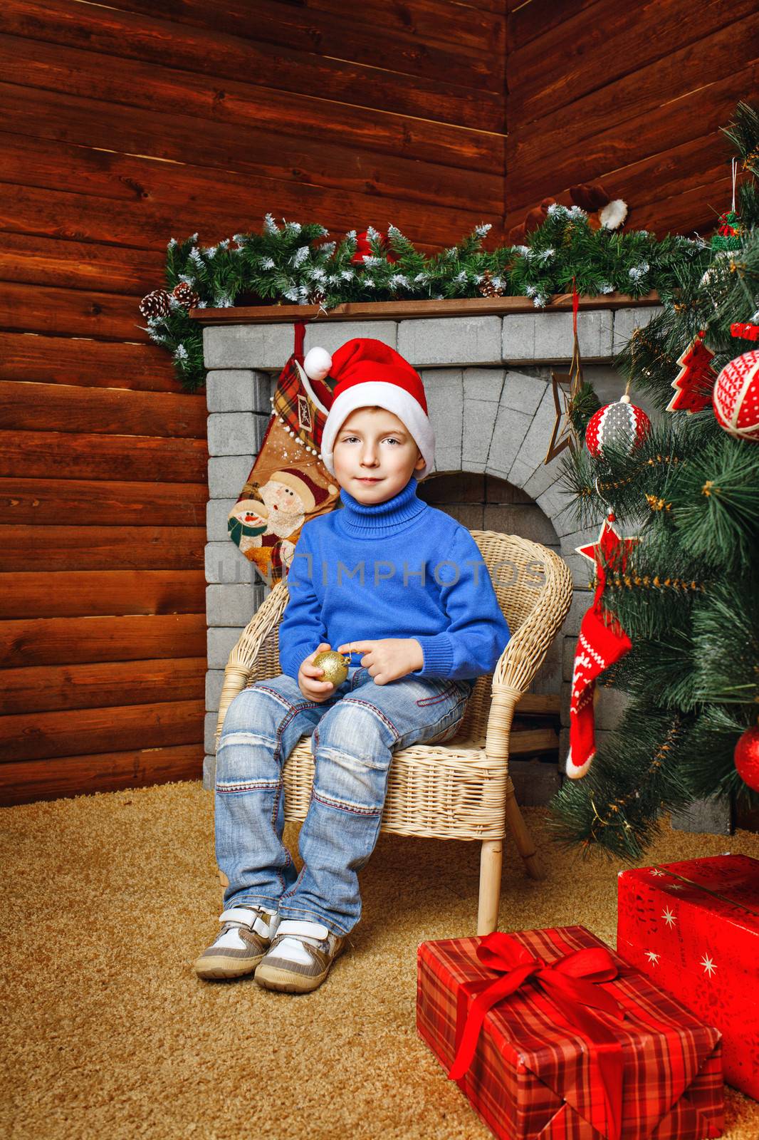 Boy in hat sitting in wicker chair near Christmas tree and gifts