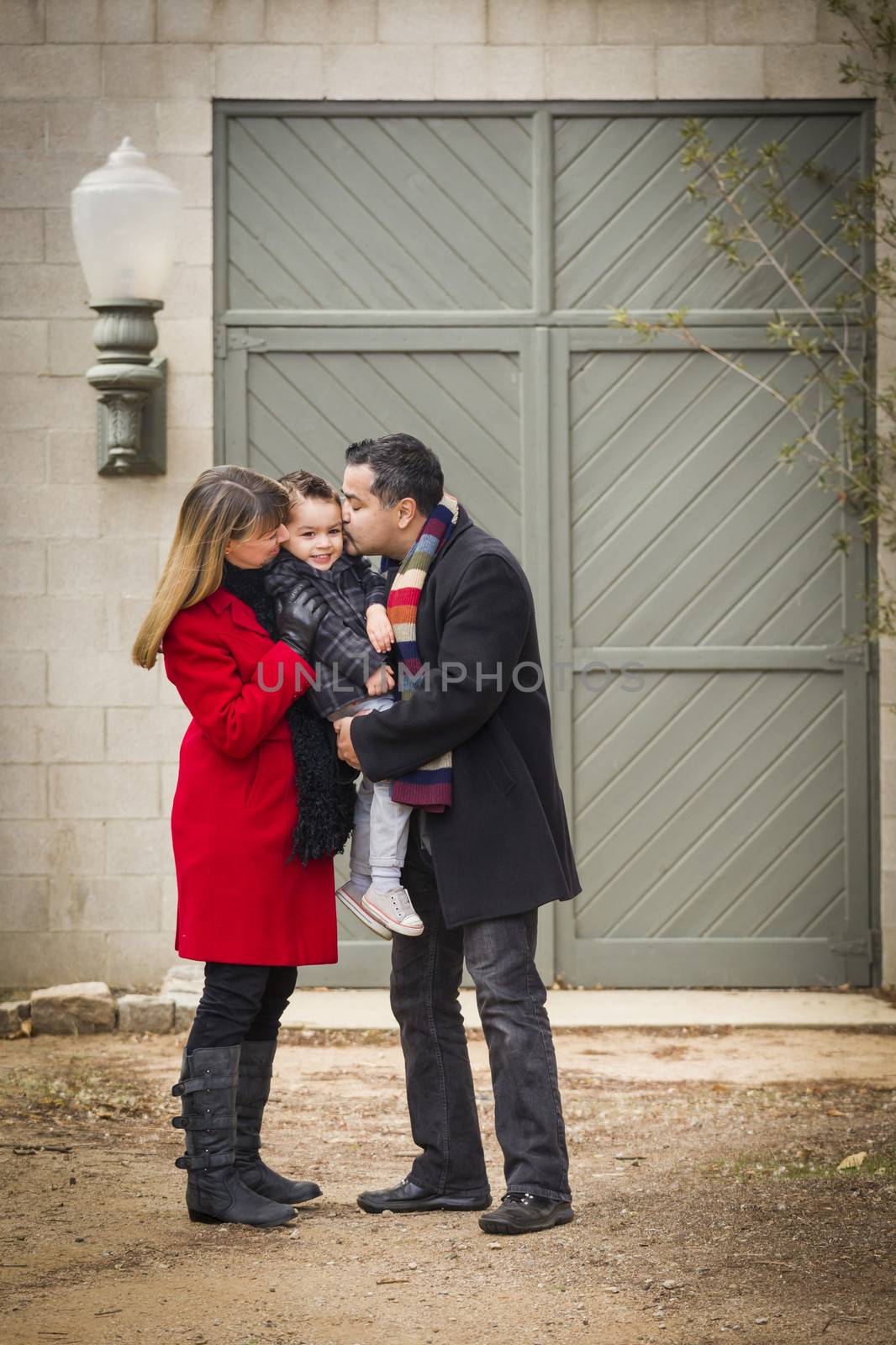 Young Mixed Race Couple in Winter Clothing Hugging and Kissing Son in Front of Rustic Building Together.