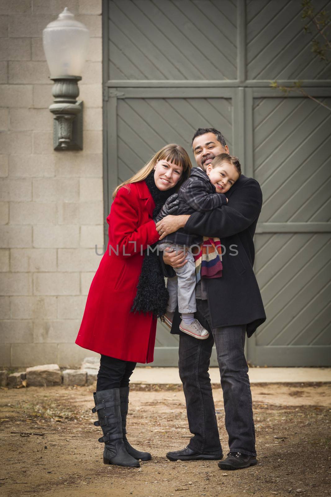 Young Mixed Race Couple in Winter Clothing Hugging and Kissing Son in Front of Rustic Building Together.