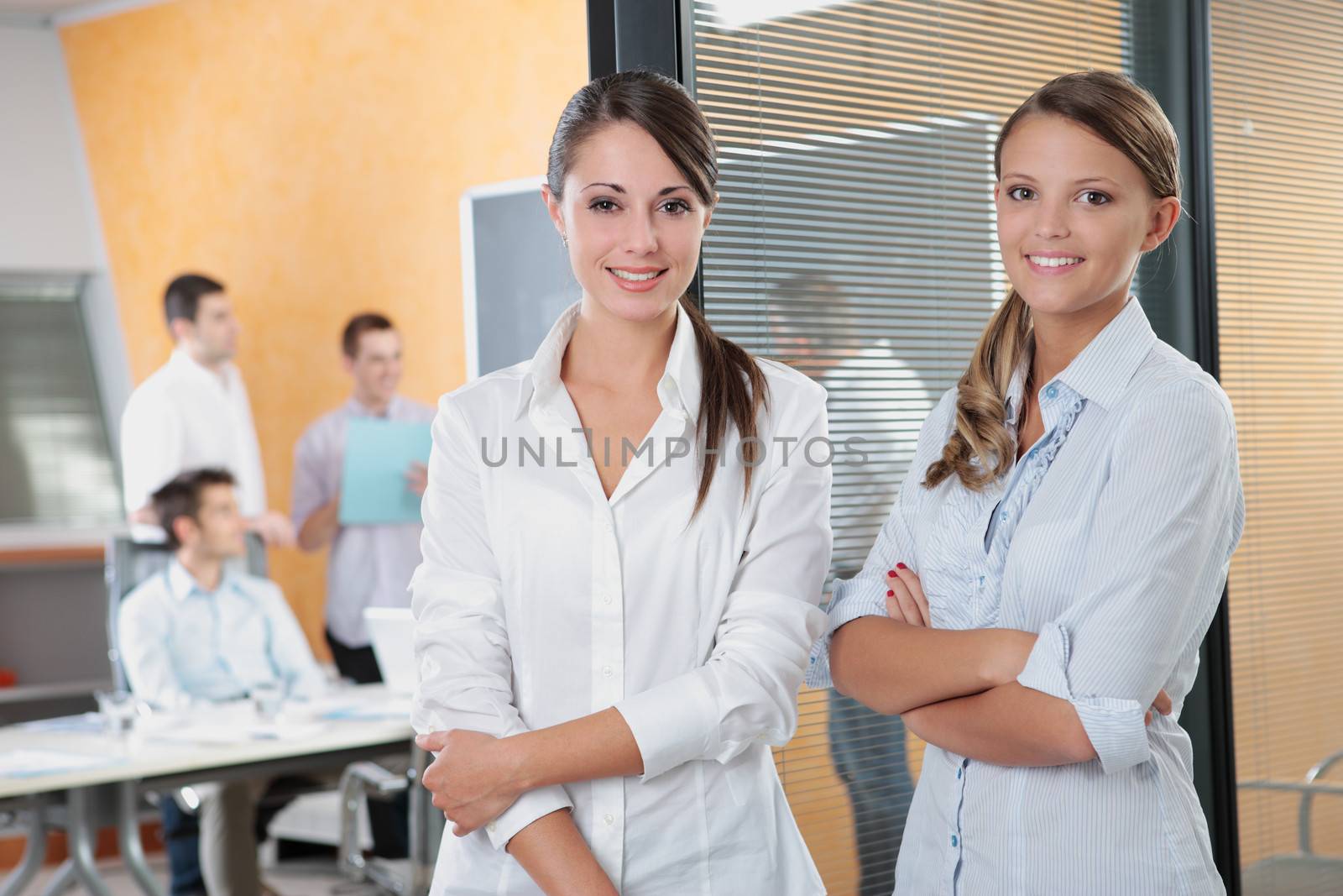 Portrait of two young business woman standing while his team talks in the background 