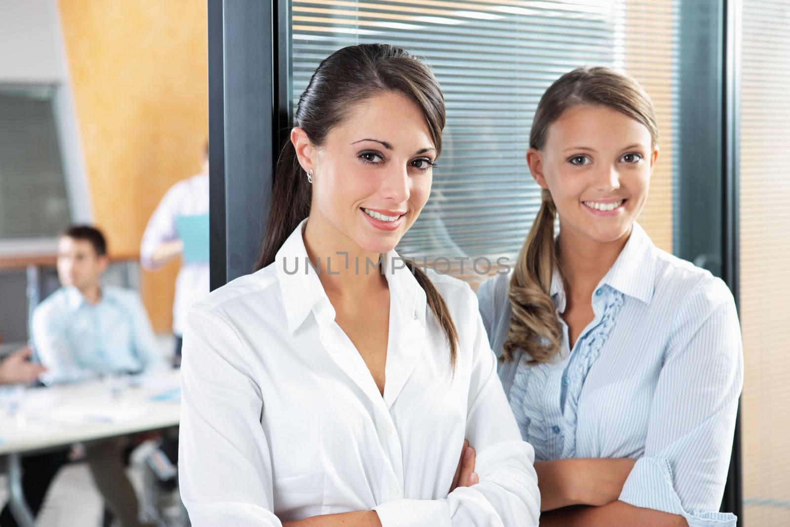 Portrait of two young business woman standing while his team talks in the background 