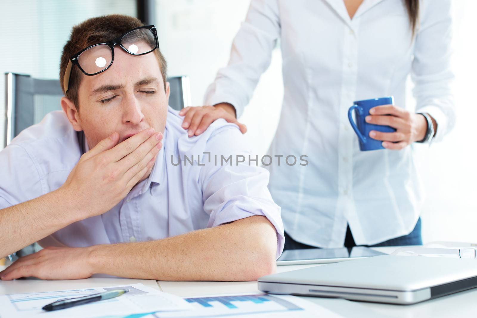Young businessman is yawning, colleague gives him a cup of coffee