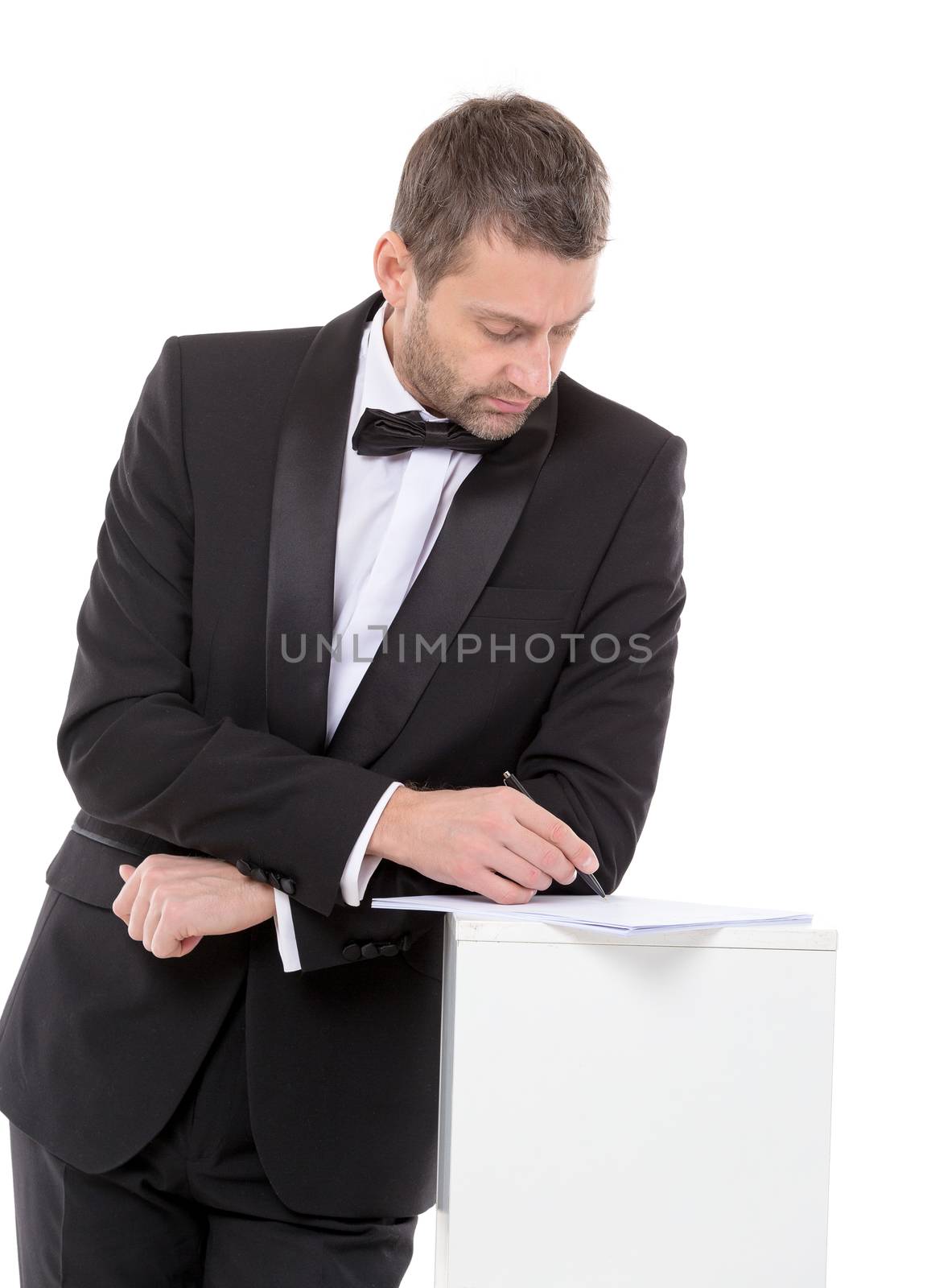 Stylish middle-aged man in a bow tie and suit standing at a pedestal completing a form pausing to read the document with a pen in his hand