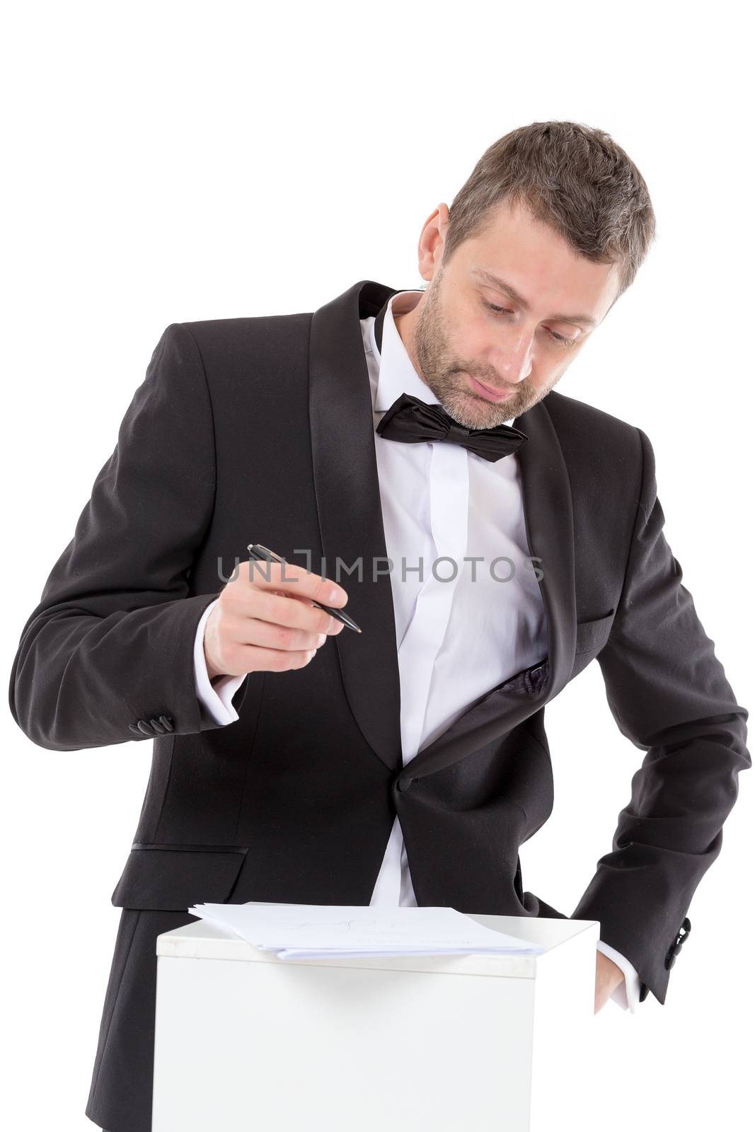 Stylish middle-aged man in a bow tie and suit standing at a pedestal completing a form pausing to read the document with a pen in his hand