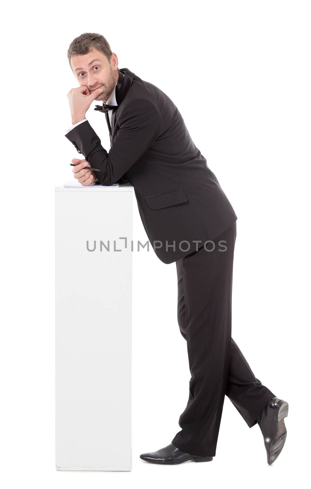 Elegant slender man in a suit and bow tie leaning nonchalantly on a white pedestal with a quizzical charismatic expression