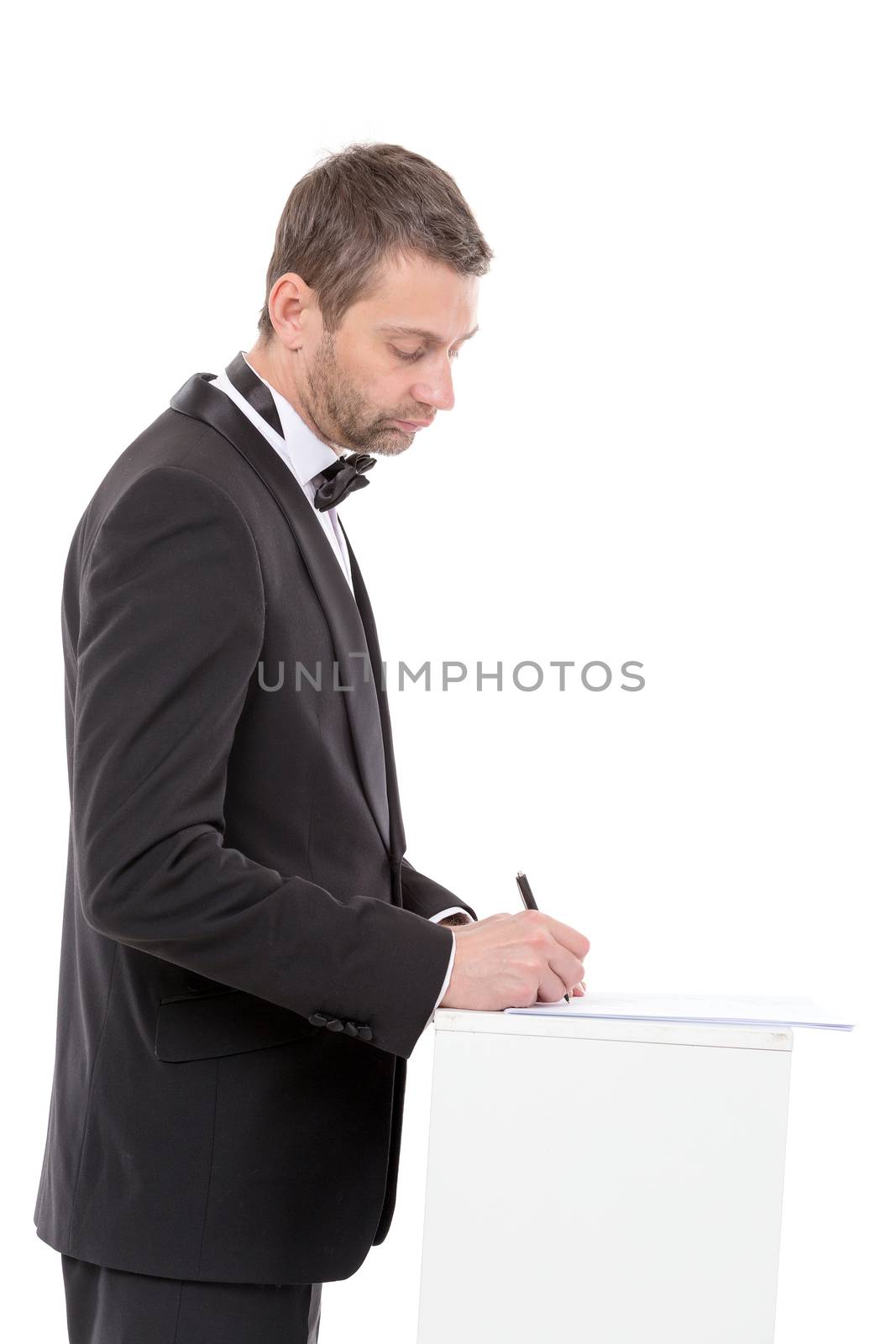Stylish middle-aged man in a bow tie and suit standing at a pedestal completing a form pausing to read the document with a pen in his hand