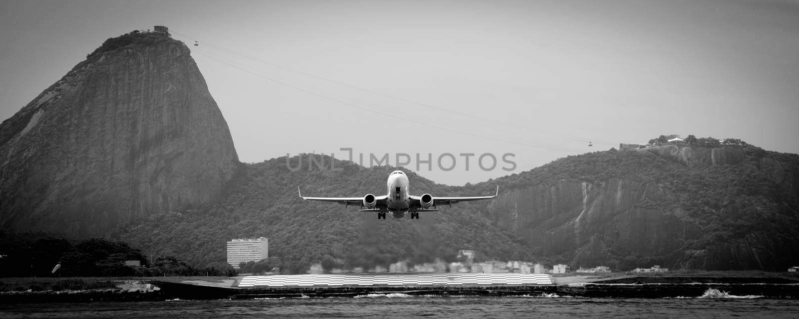 Black and white view of aircraft taking off over Guanbara Bay with Rio de Janiero and Sugar Loaf mountain in background, Brazil.