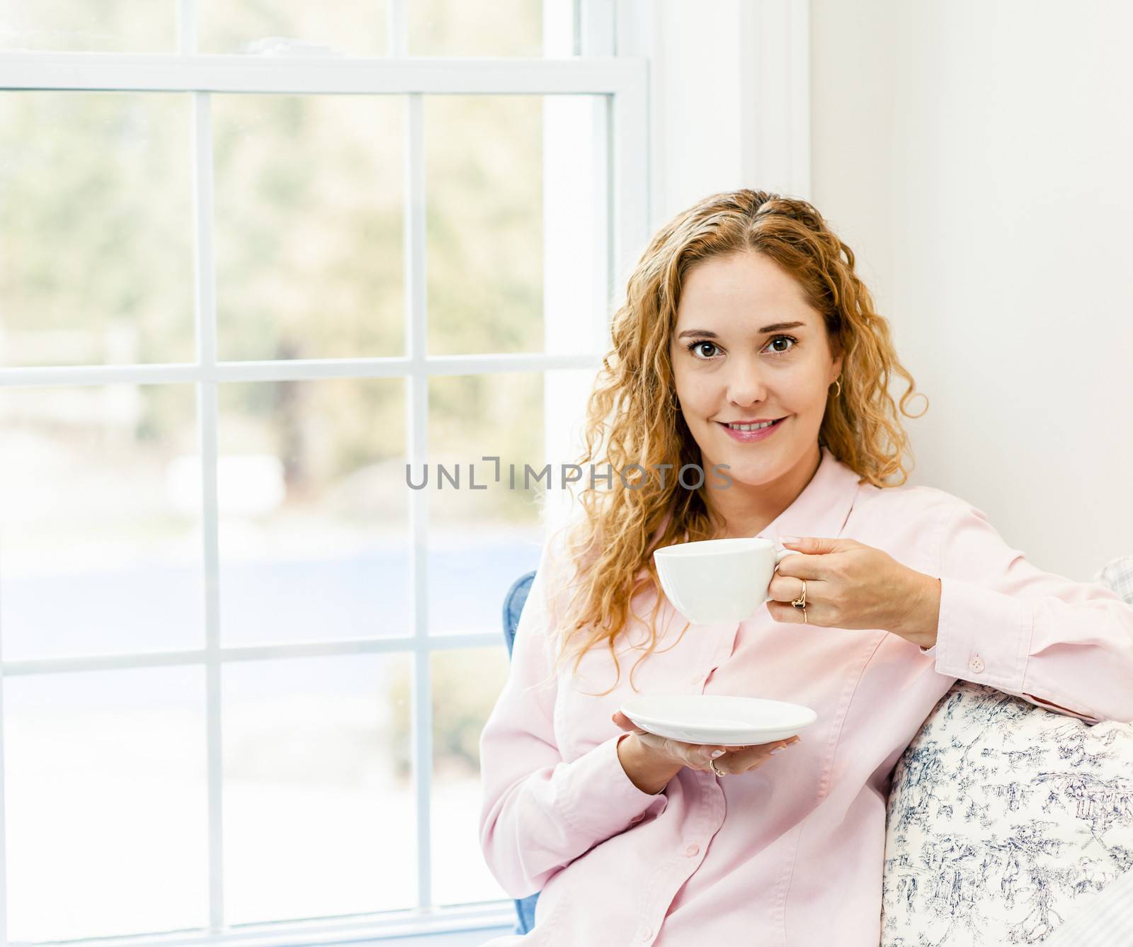 Smiling caucasian woman relaxing by window holding cup of coffee