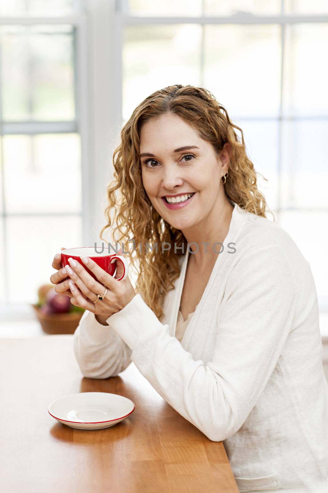 Portrait of smiling woman holding red coffee cup sitting at table in home kitchen by window