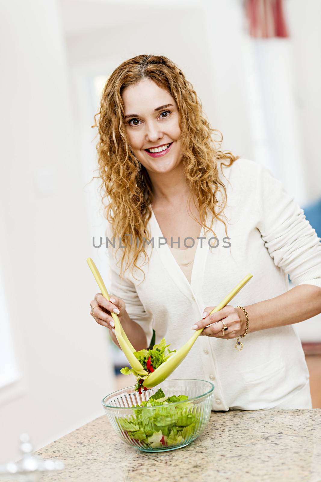 Smiling woman tossing salad in kitchen by elenathewise