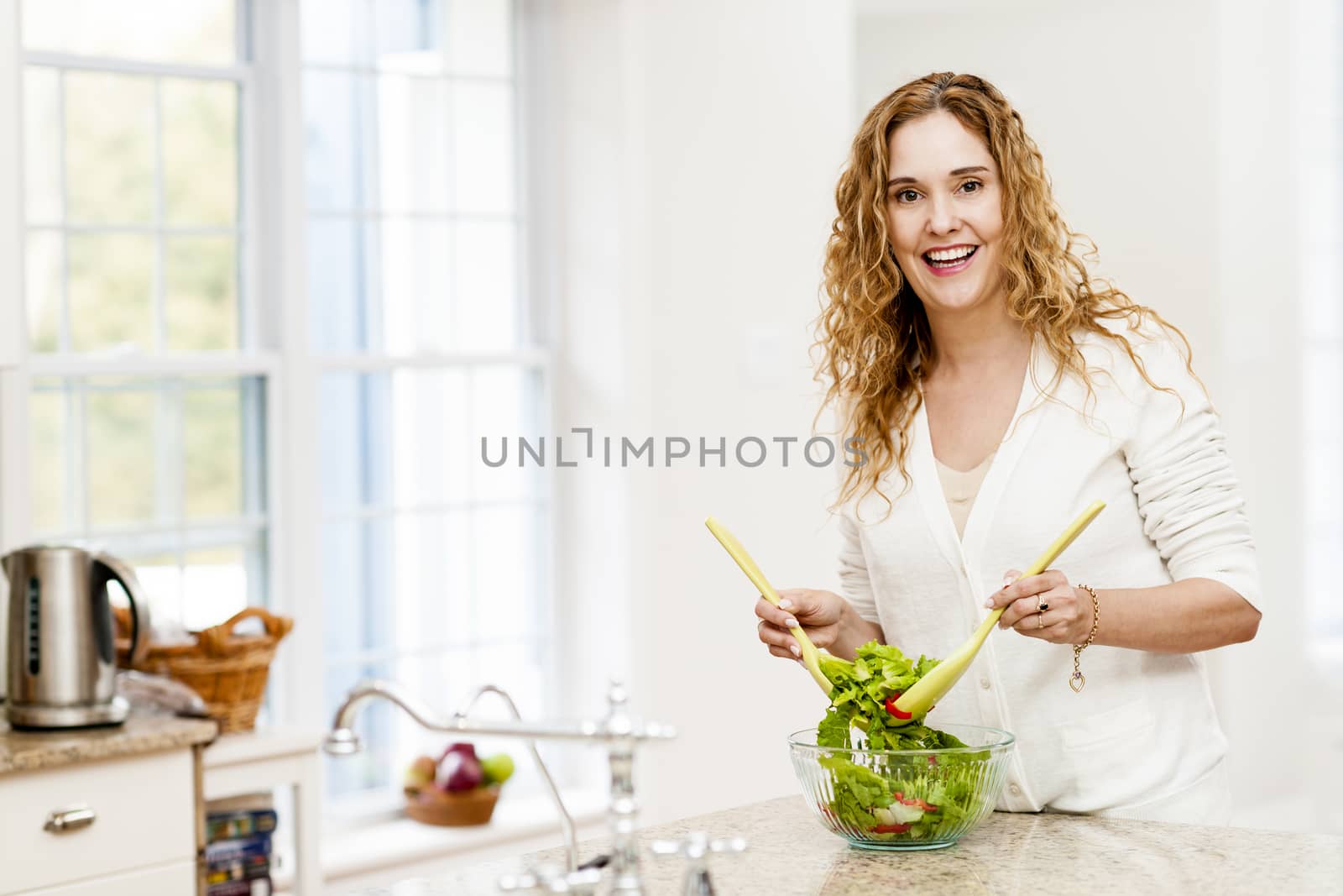 Smiling woman tossing salad in kitchen by elenathewise