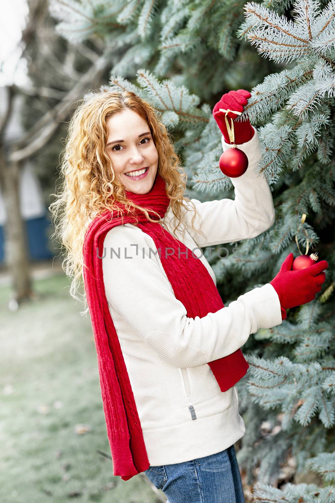 Joyful woman hanging Christmas ornaments on spruce tree outdoors in yard near home