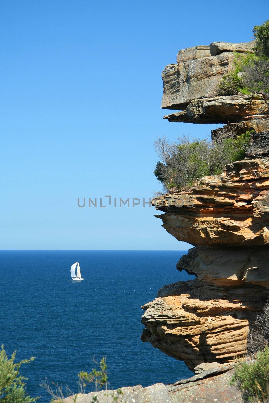 Rocky sandstone cliffs along the coast of Sydney, Australia with a white sailboat in the distance.