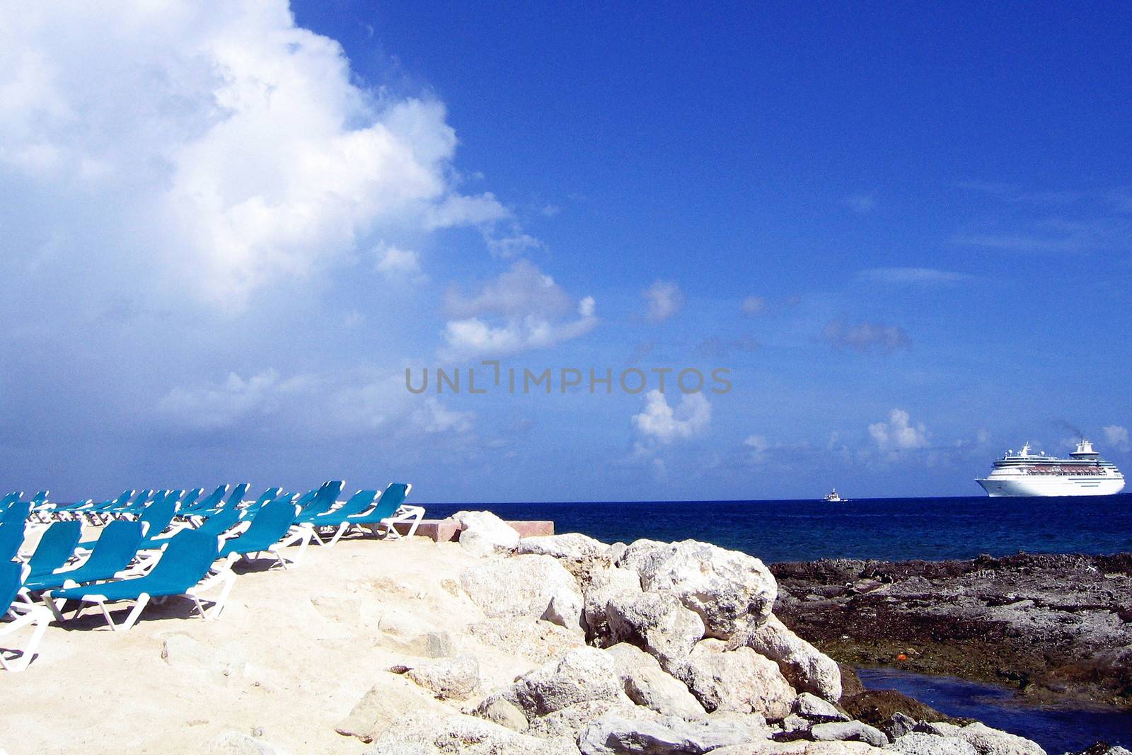 Empty beach chairs on rocky coastline with cruise ship sailing in blue sea background.
