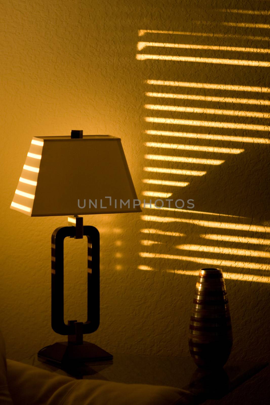 Bright sunshine in a dark room casts lines through window blinds and on to a lamp.