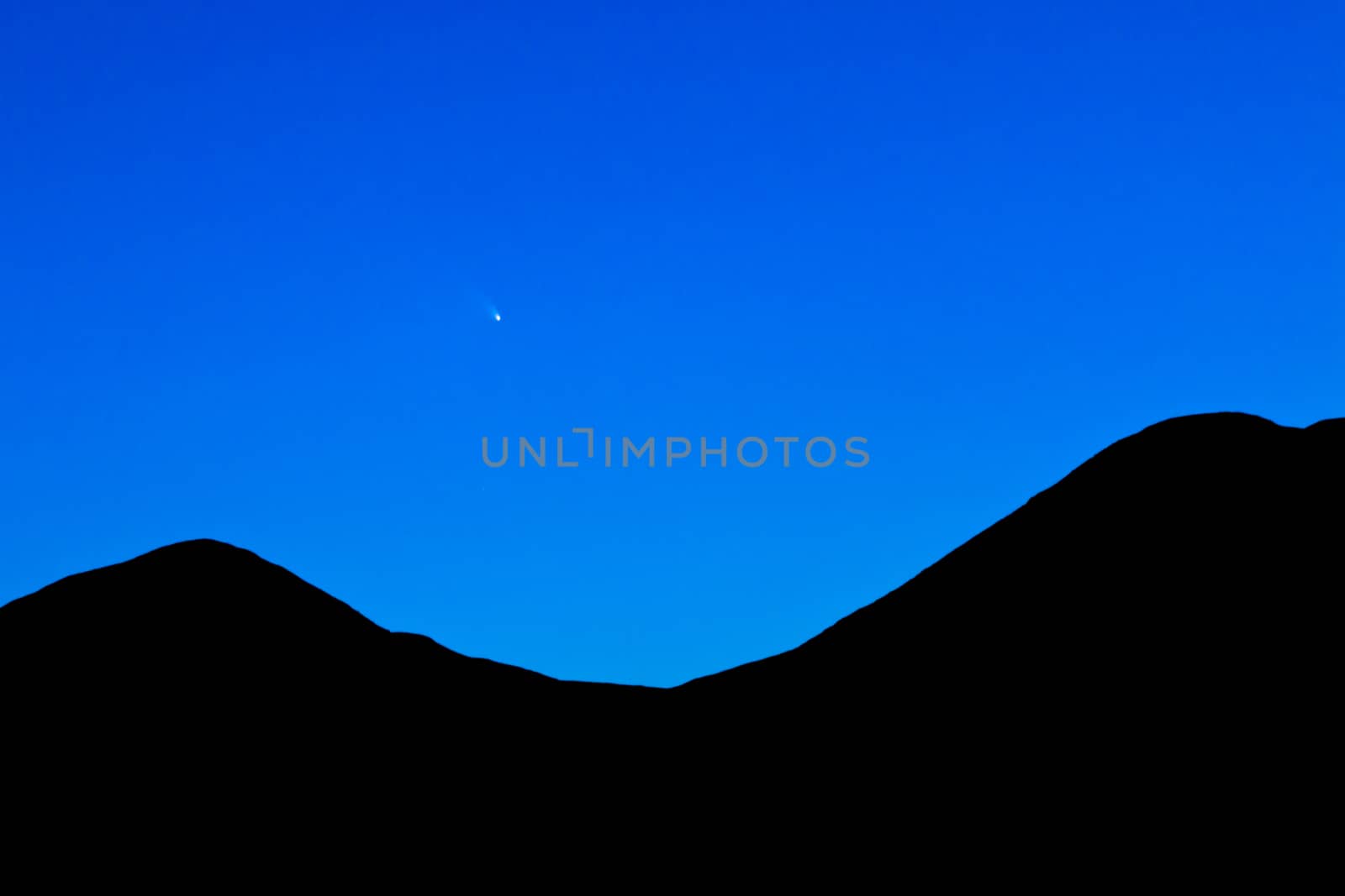 Comet C/2011 L4 PanSTARRS over silhouette of mountainous horizon on blue western night sky just after sunset