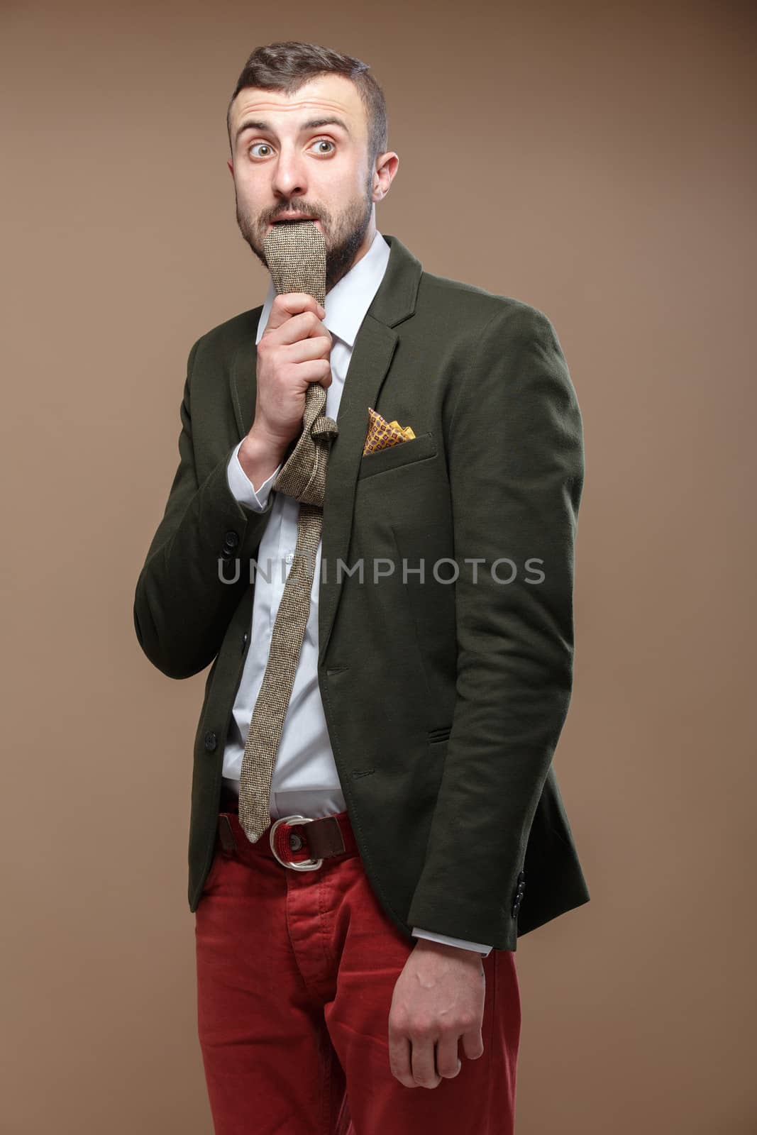 Young stylish man in a green suit and orange jeans with a tie in the mouth. Beige background.