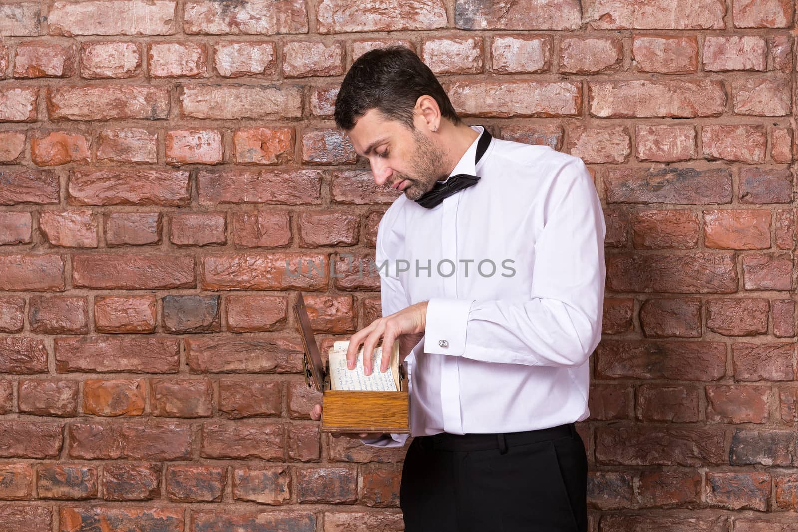 Man standing reading a old document from a wooden box which he is holding in his hand while standing in front of a brick wall