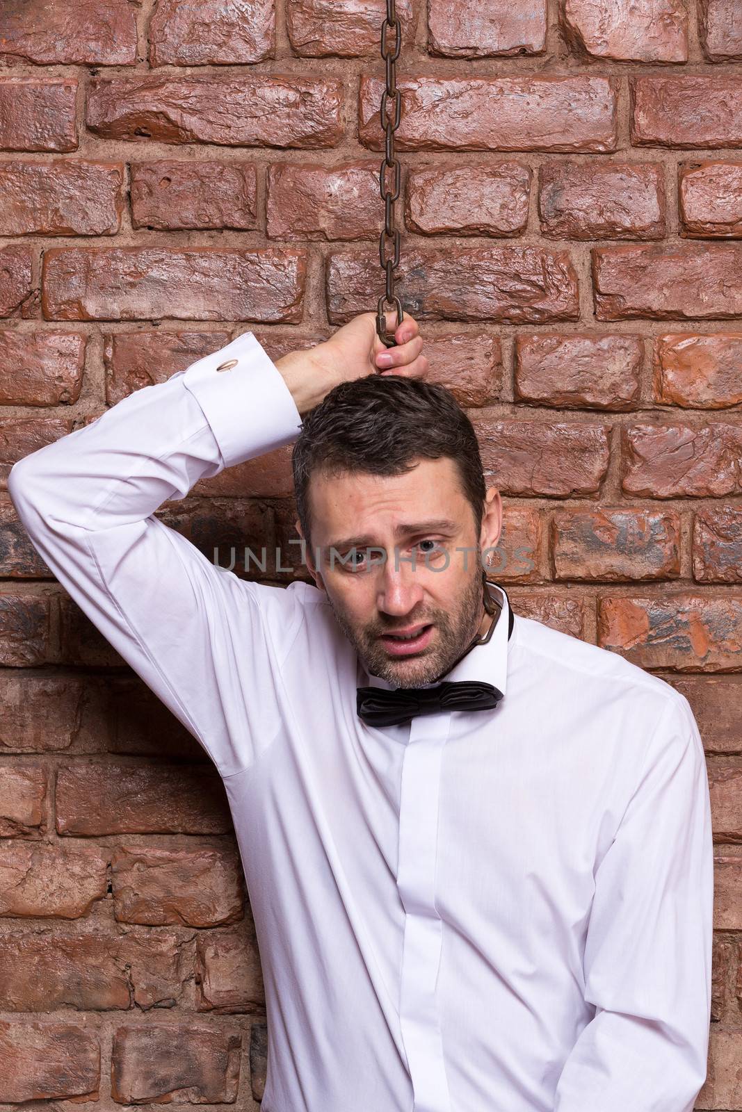 Conceptual image of a desperate handsome businessman in a bow tie standing against a brick wall with a chain around his neck and a disconsolate depressed expression