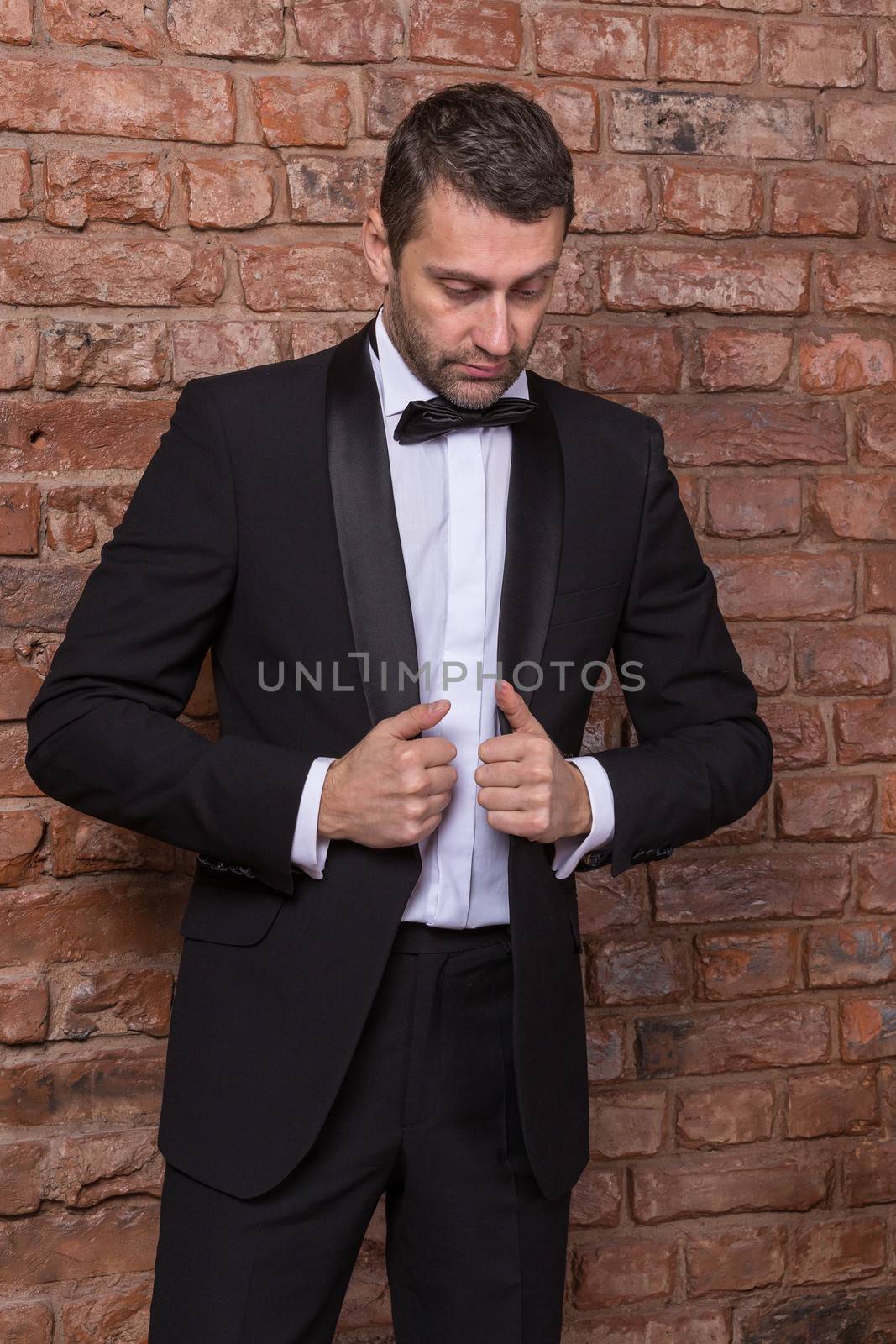Elegant handsome macho man in a bow tie and tuxedo leaning against a brick wall giving the camera a sultry look