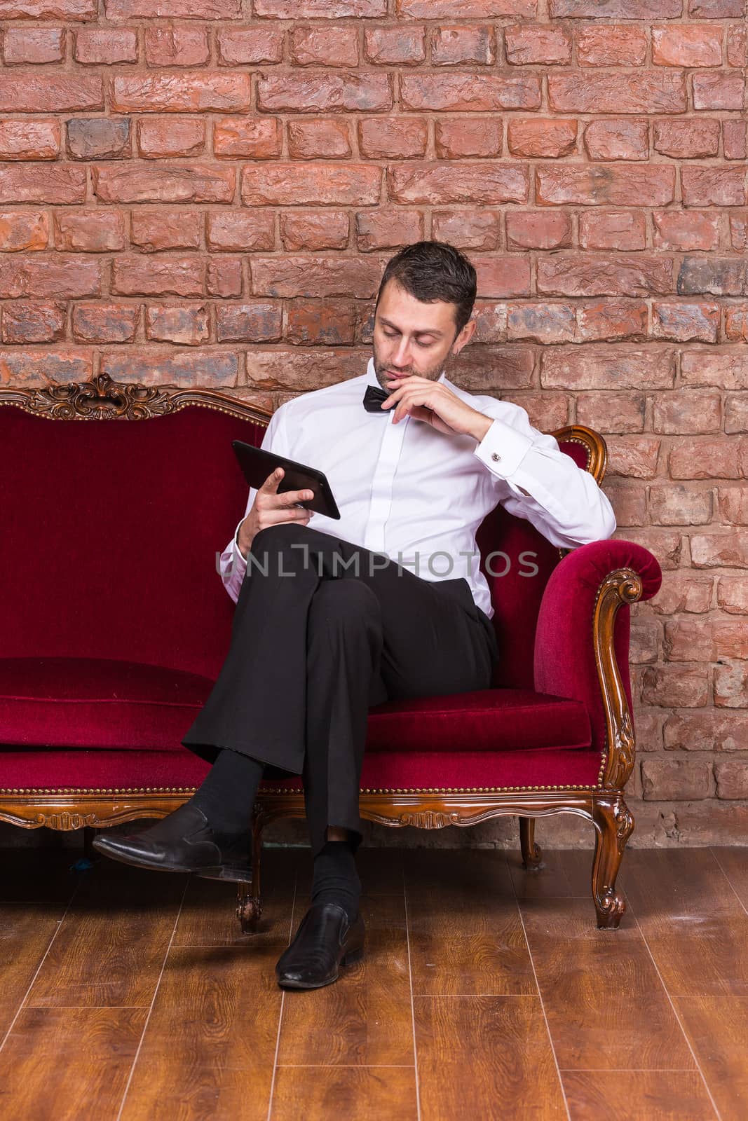 Conceptual image of an elegant businessman lying relaxing on a settee against a brick wall and reading tablet