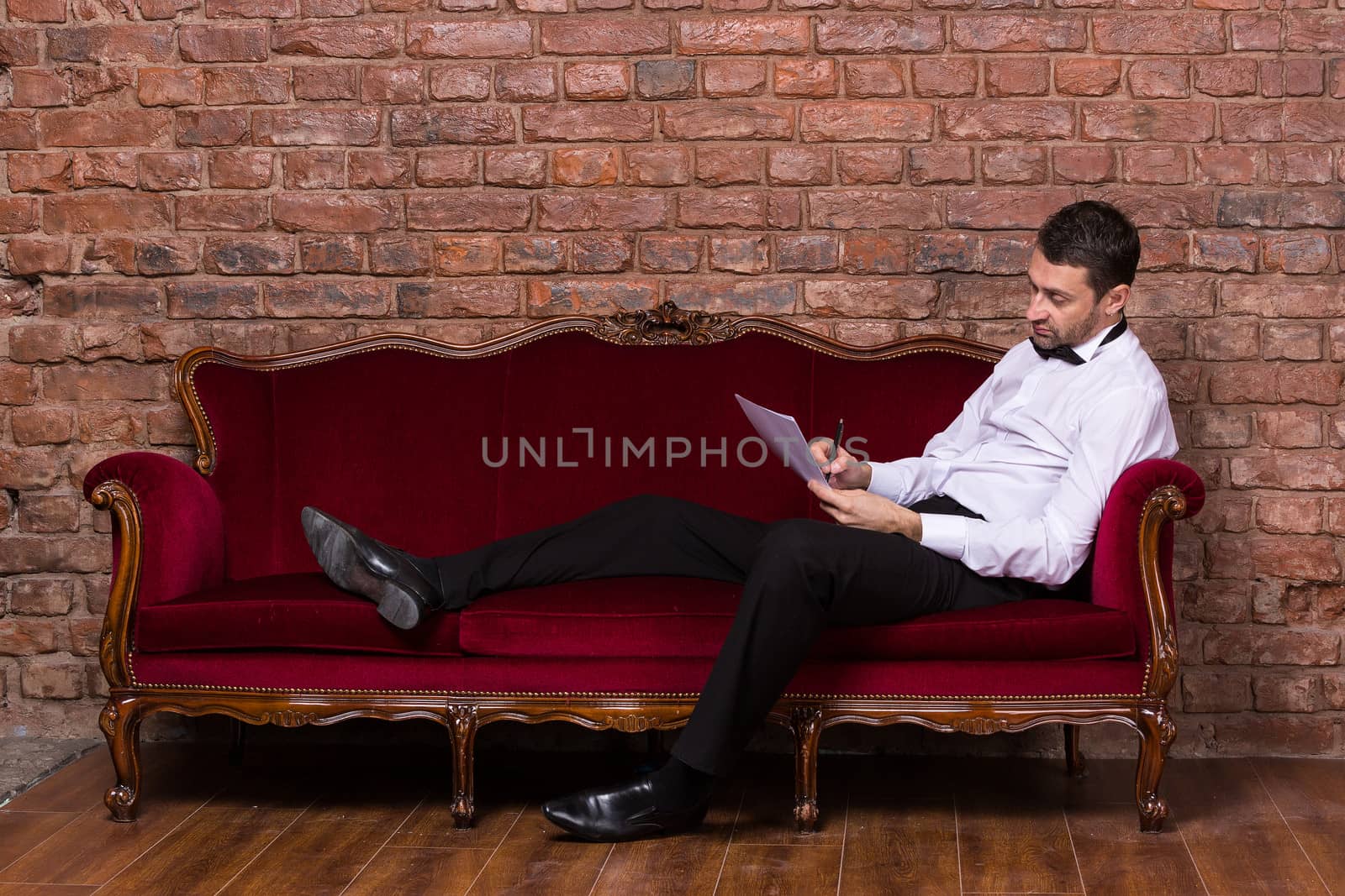 Conceptual image of an elegant businessman lying relaxing on a settee against a brick wall and reading paperwork