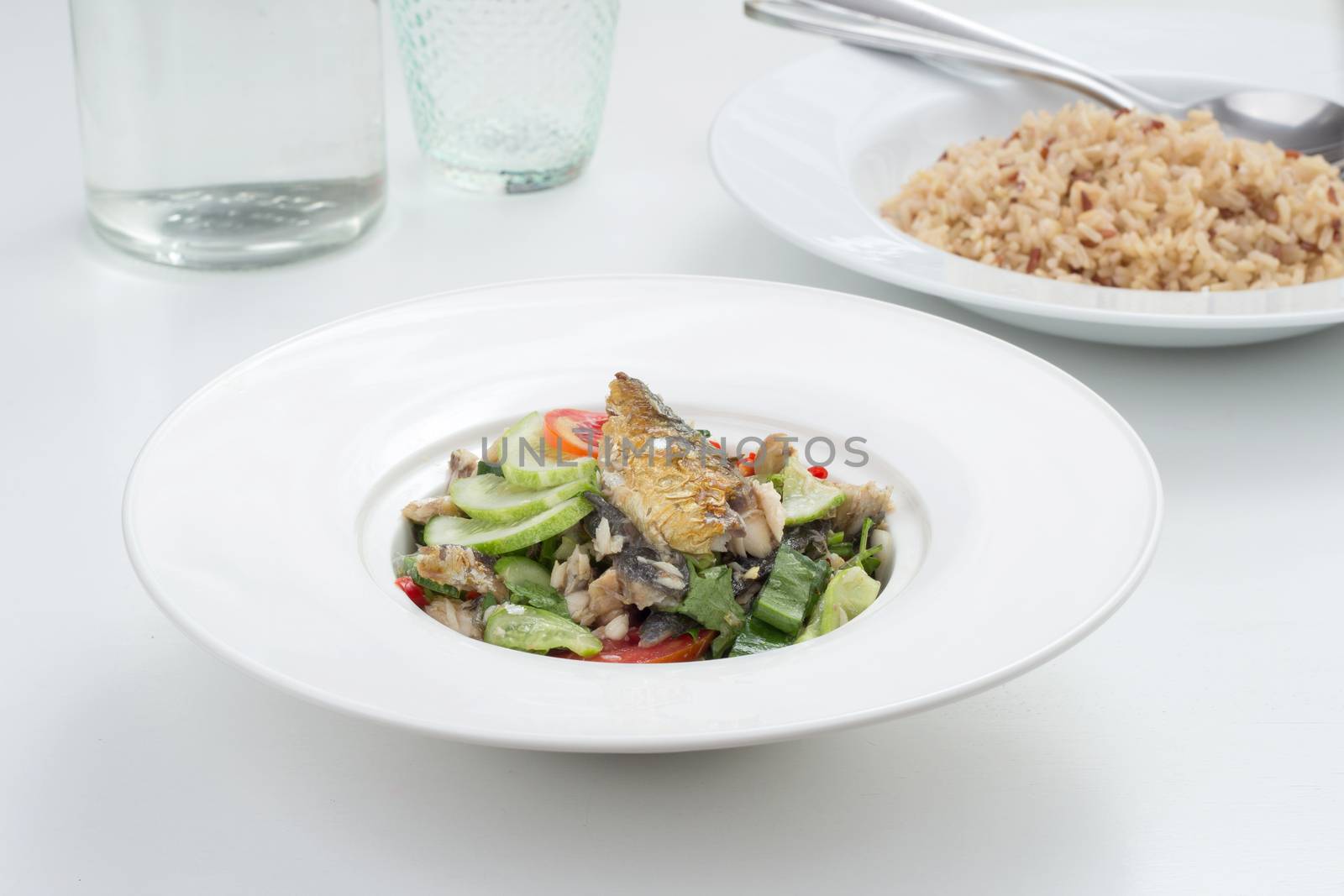 Thai appetizer - spicy mackerel salad with lemon grass, shallots, raw mango, chili and lettuce on a white plate. 
unpolished rice on the back