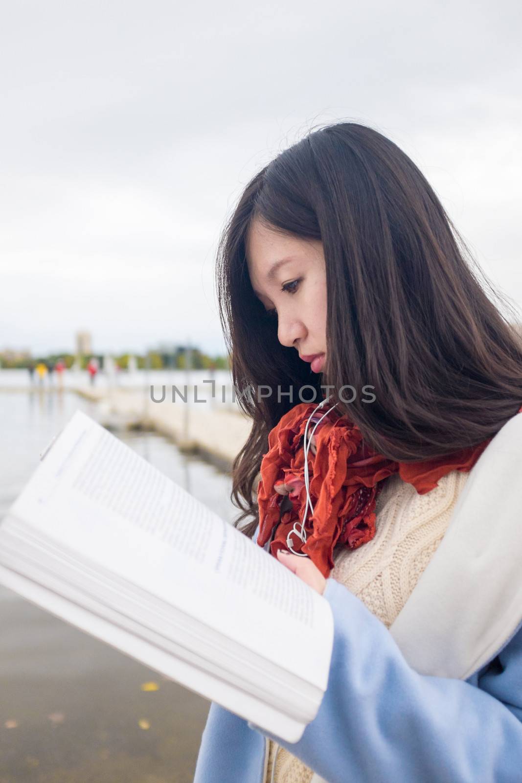 Attractive Asian girl in blue coat reading a book near a lake